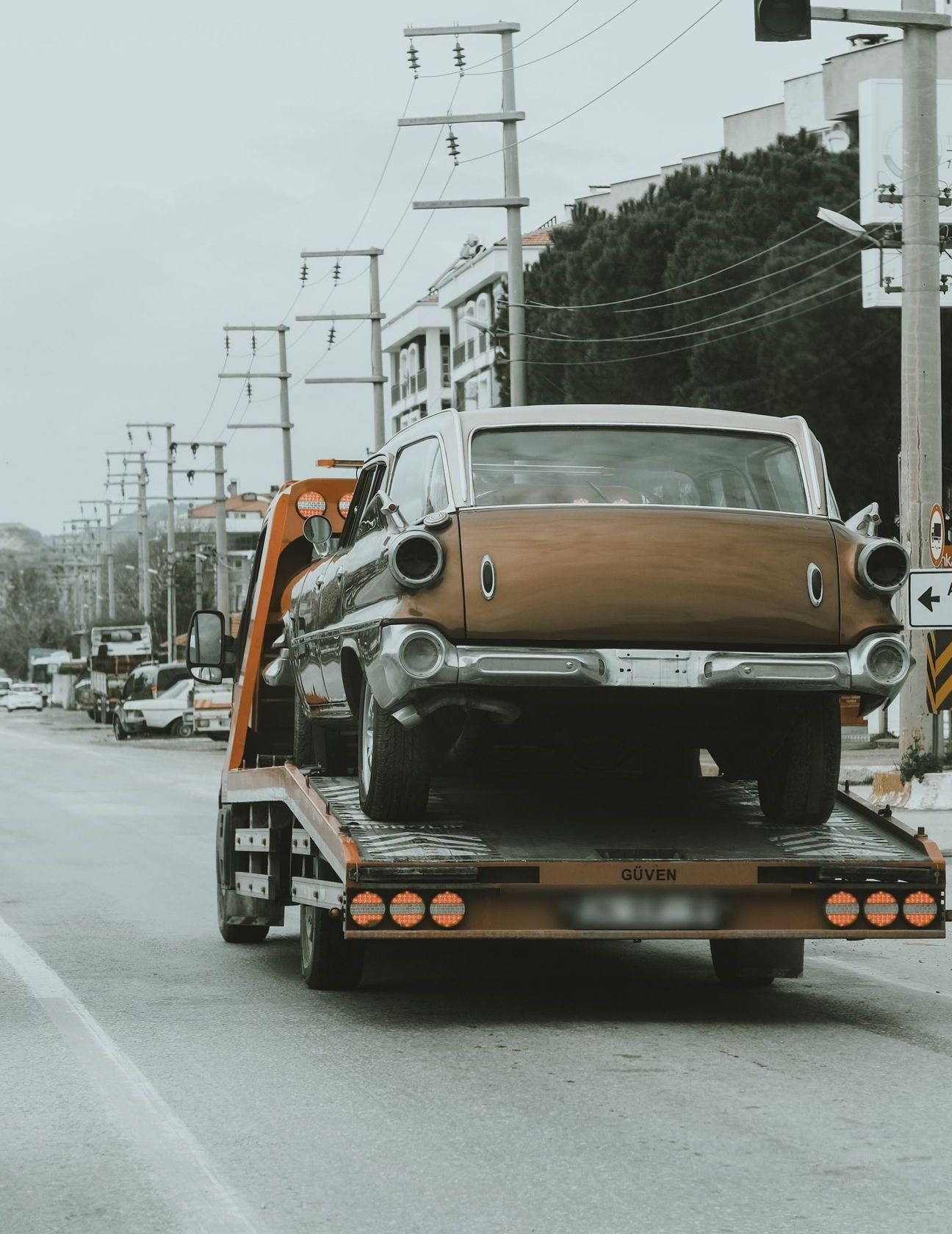 A vintage car is being towed through city streets on a tow truck, showcasing an urban environment.